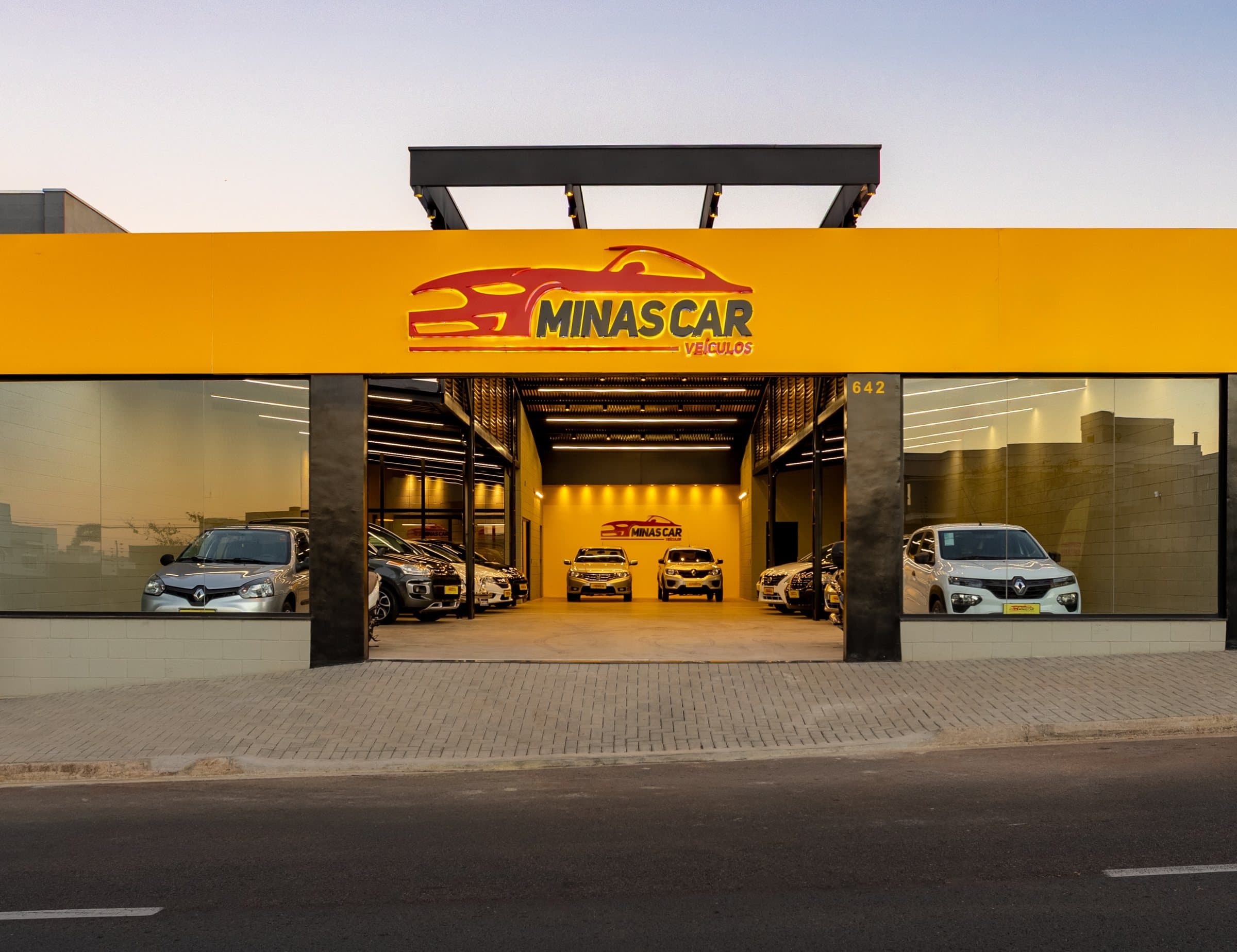 Fachada da Minas Car ao entardecer: letreiro amarelo com o logo, vitrine de vidro e os carros no salão iluminado