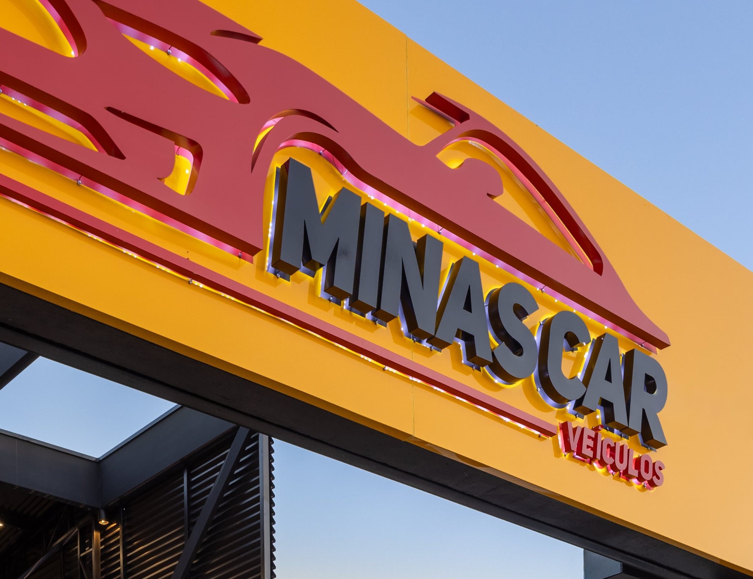 Letreiro da Minas Car Veículos de perto, com o logo vermelho e as letras pretas sobre o amarelo