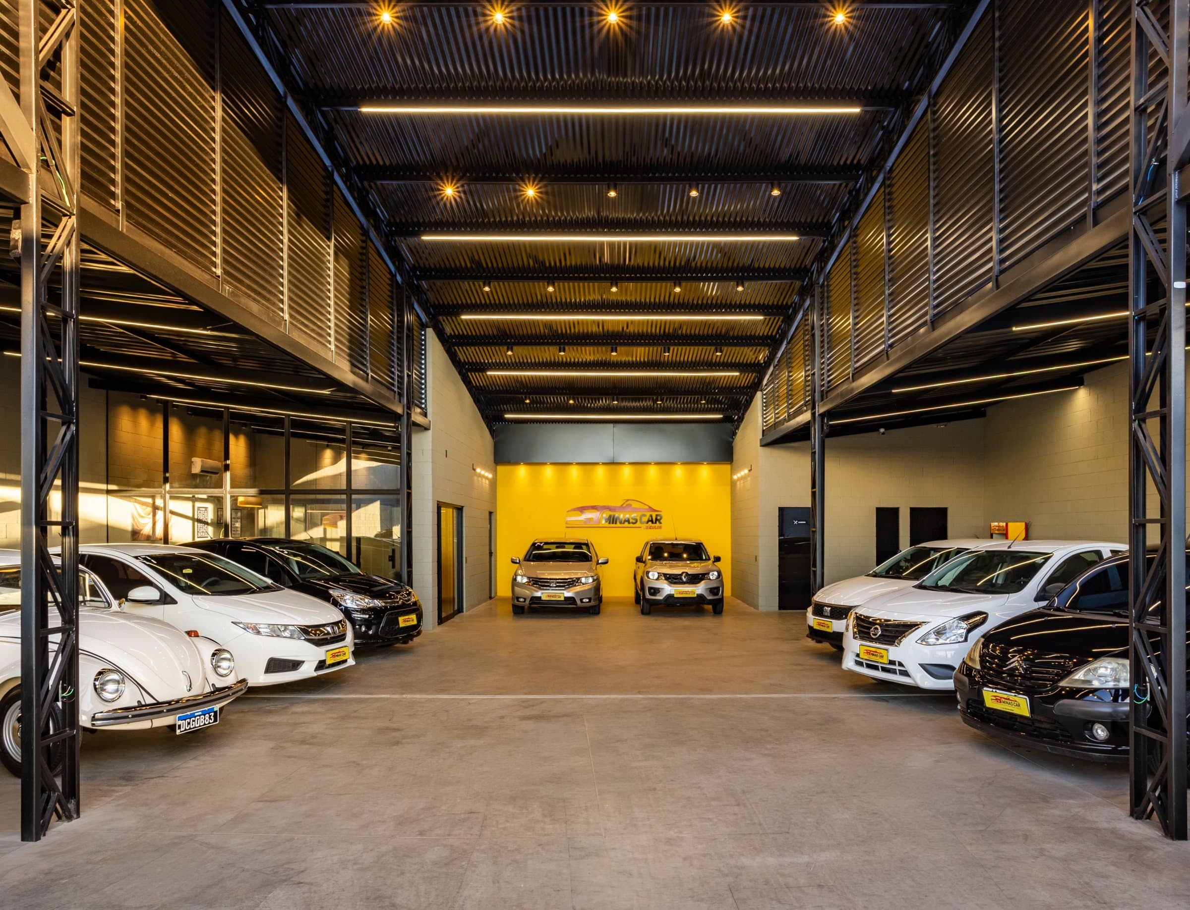 O salão da Minas Car por dentro: corredor com estrutura metálica preta, luzes no teto, carros dos dois lados e a parede amarela do logo no fundo